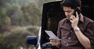 A man talks on his phone while looking at a document in front of a car with an open hood.