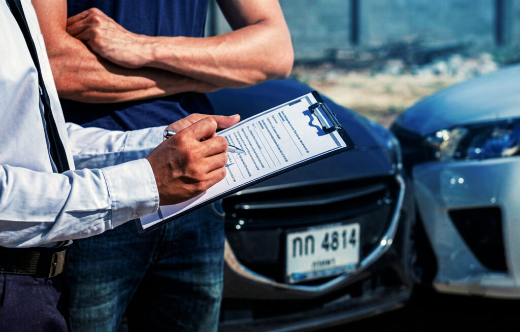 An insurance claims adjuster fills out a report on a clipboard while standing in front of two damaged cars at the scene of a vehicle accident.