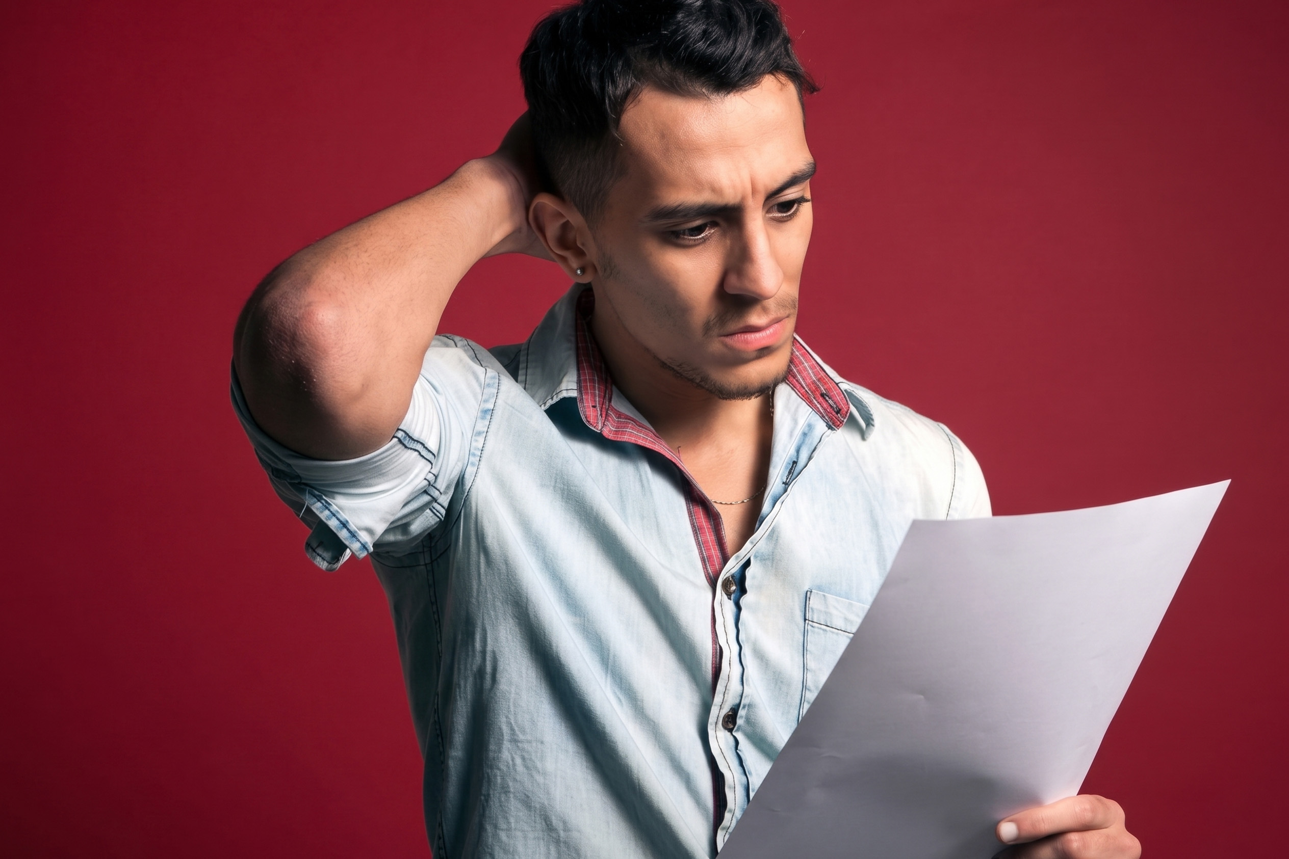 A confused man looking down at a document while scratching his head in front of a red background.