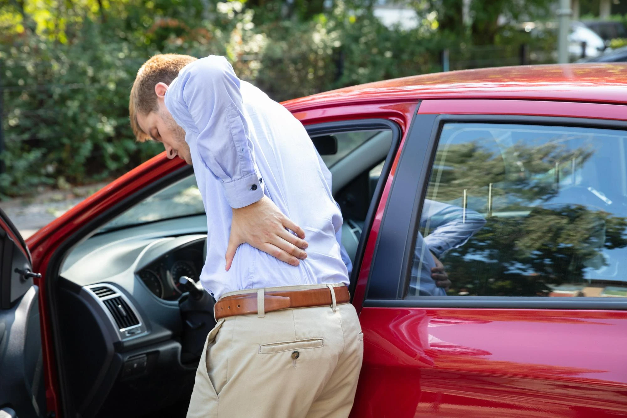 A man holding his lower back in pain while stepping out of a red car.