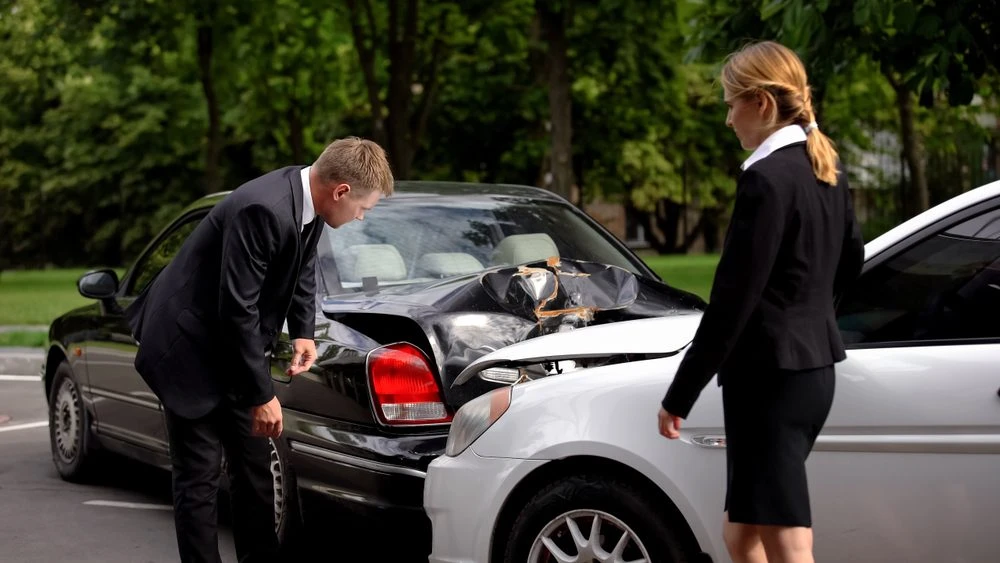 A man and a woman in business suits inspecting the damage after a rear-end car collision.