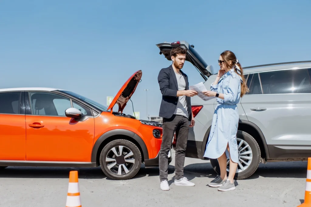 A man and a woman stand between a grey SUV and an orange hatchback, reviewing documents following a minor car accident.